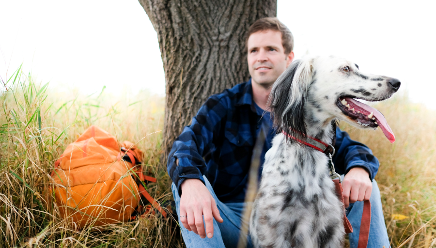 Чоловік сидить перед деревом із помаранчевим рюкзаком на траві, а його собака сидить перед ним